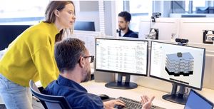 A man working at a desk with dual monitors, displaying data and a building model, while a woman in a yellow sweater looks on.