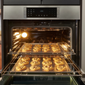 Chocolate chip cookies baking on two trays inside a lit oven, with a digital control panel above.