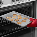 A hand wearing a red oven mitt places a tray of freshly baked chocolate chip cookies into an open oven.