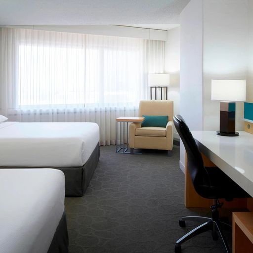 Modern hotel room with two beds, a desk, chair, lamps, and large window with sheer curtains, creating a bright and welcoming atmosphere.