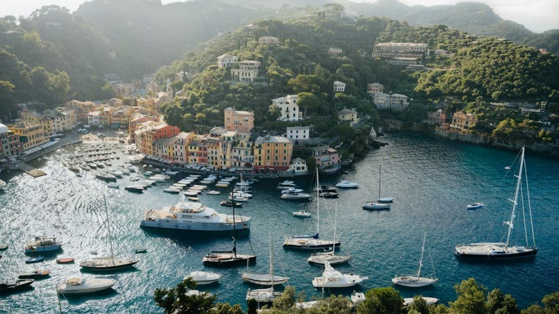 Scenic view of Portofino harbor with vibrant buildings, lush hills, and numerous boats anchored in the blue-green water under a hazy sky.