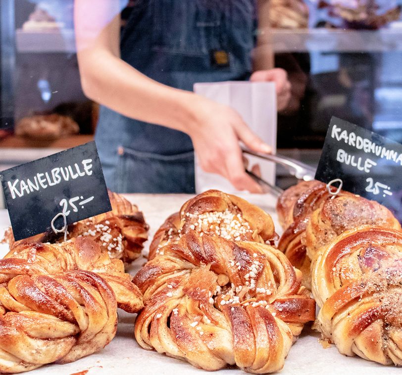 A bakery display of Swedish cardamom and cinnamon buns, with a person using tongs behind a glass. Price tags read "25 kr" for each type.