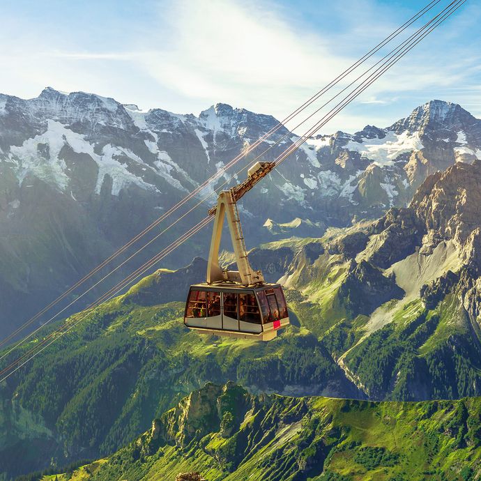 A cable car ascending through rugged, green mountain terrain with snow-capped peaks in the background under a clear blue sky.