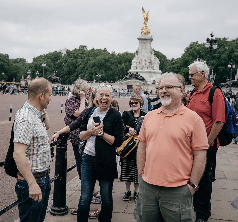 A group of people smiling and chatting near a monument with a golden statue on top, surrounded by greenery and a cloudy sky.