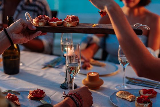 People dining outdoors, sharing a tray of appetizers, with wine glasses on the table and a pool in the background.