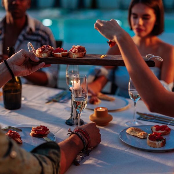 People dining outdoors, sharing a tray of appetizers, with wine glasses on the table and a pool in the background.