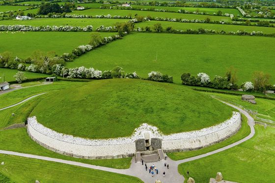 Aerial view of Newgrange, a large circular mound with a stone entrance, surrounded by lush green fields and scattered trees under a cloudy sky.