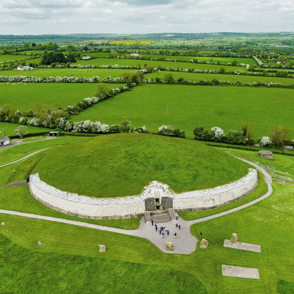 Aerial view of Newgrange, a large circular mound with a stone entrance, surrounded by lush green fields and scattered trees under a cloudy sky.