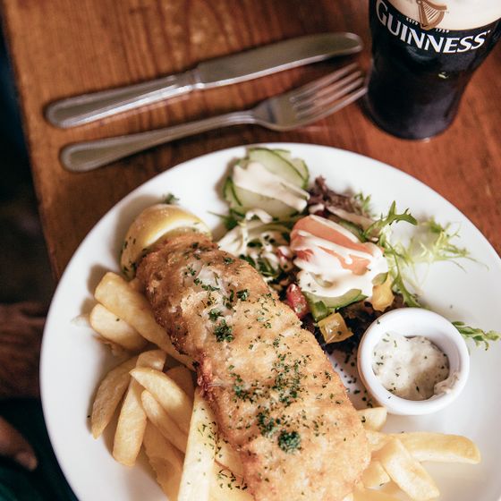 Plate of fish and chips with tartar sauce, lemon wedge, and salad. Knife, fork, and a pint of Guinness on a wooden table.