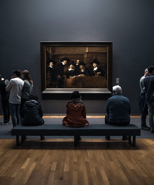 People observing a large framed painting in an art gallery, with some seated and others standing, all facing the artwork on a dark wall.