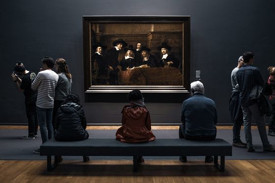 People observing a large framed painting in an art gallery, with some seated and others standing, all facing the artwork on a dark wall.
