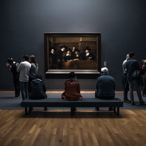 People observing a large framed painting in an art gallery, with some seated and others standing, all facing the artwork on a dark wall.