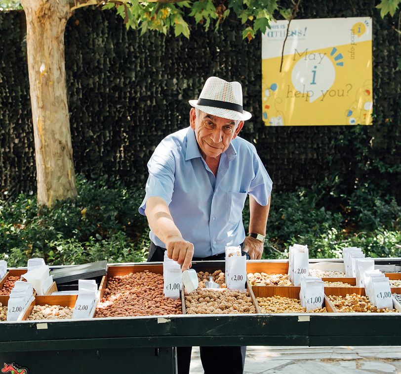 Man in a hat selling nuts at an outdoor stall, surrounded by greenery. Various nuts are displayed with price tags.
