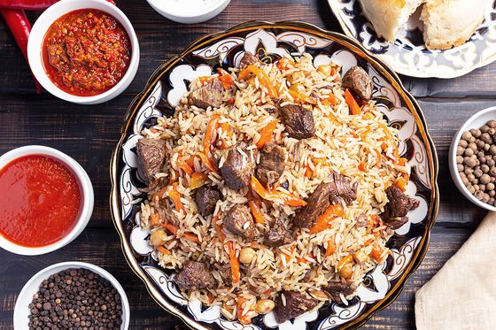 A plate of rice pilaf with meat and vegetables, surrounded by bowls of sauces, spices, garlic, and a dish of pastries on a wooden table.