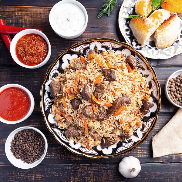 A plate of rice pilaf with meat and vegetables, surrounded by bowls of sauces, spices, garlic, and a dish of pastries on a wooden table.