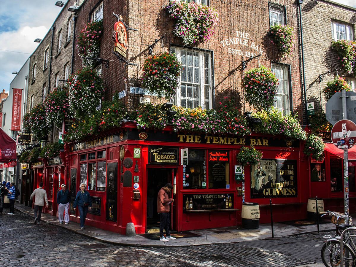 A vibrant pub with red facade and hanging flower baskets, set on a cobblestone street under a partly cloudy sky.