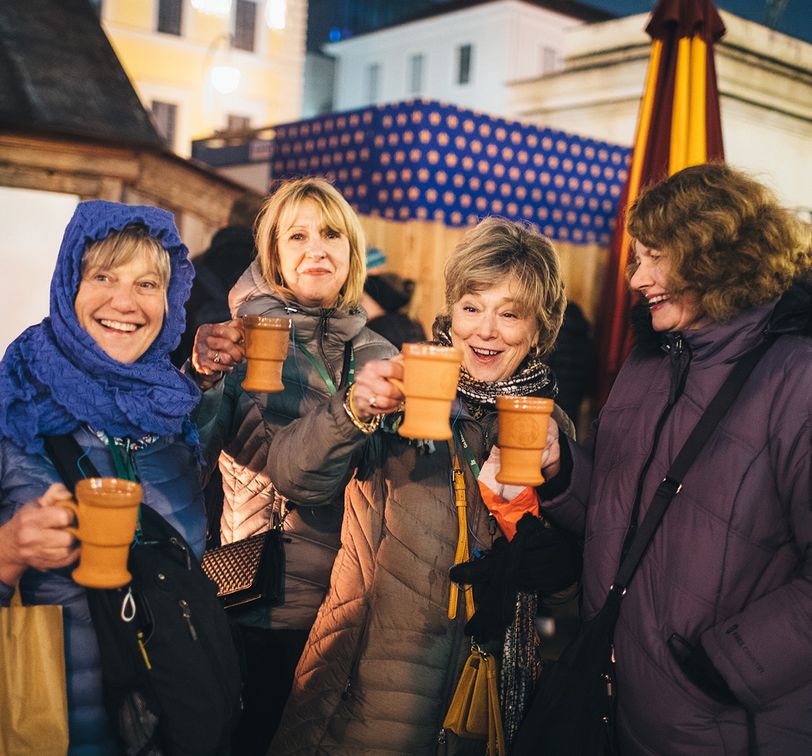 Four women in warm coats smile and toast with ceramic mugs at an outdoor festive market, surrounded by illuminated booths and decorations.