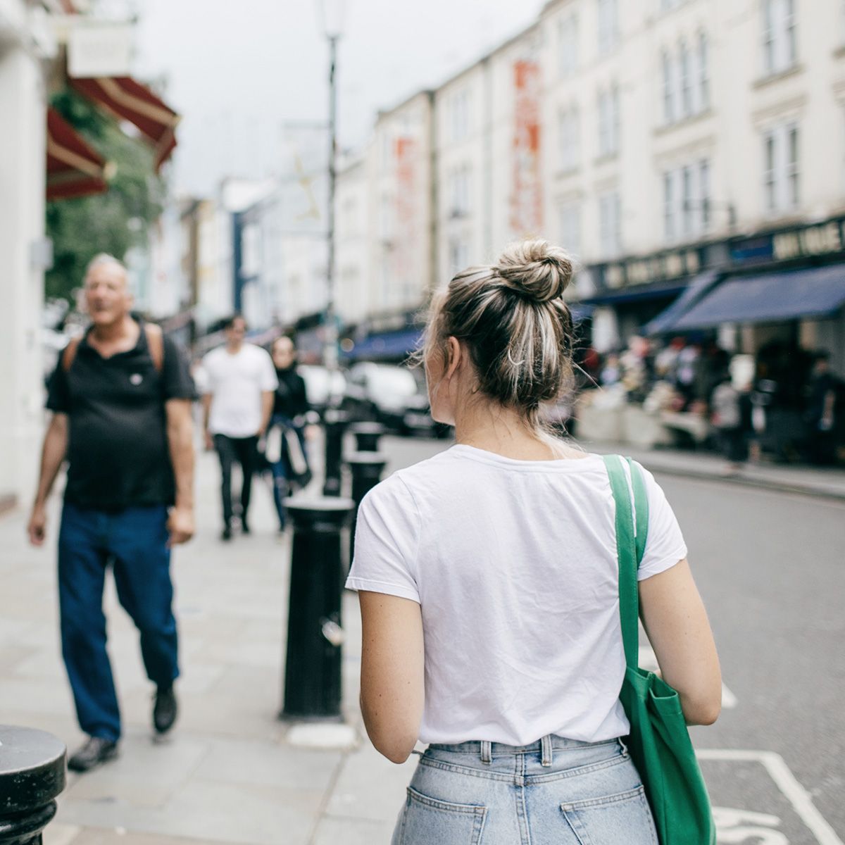 Person with a bun walks down a busy street, wearing a white shirt and carrying a green bag, with shops and pedestrians in the background.