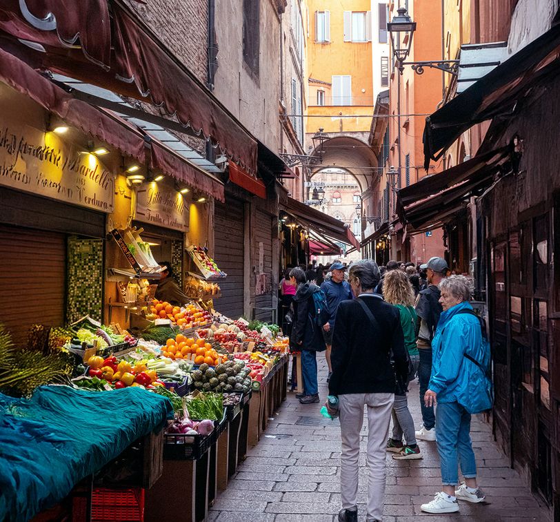 People browse a street market with colorful produce under awnings in a narrow, cobblestone alley surrounded by aged buildings.