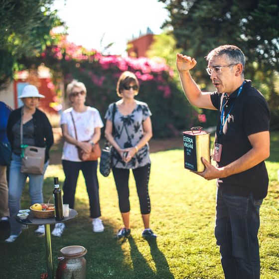 A man demonstrates olive oil to a group outdoors. A table with bottles and fruits is nearby. The group listens attentively on a sunny day.