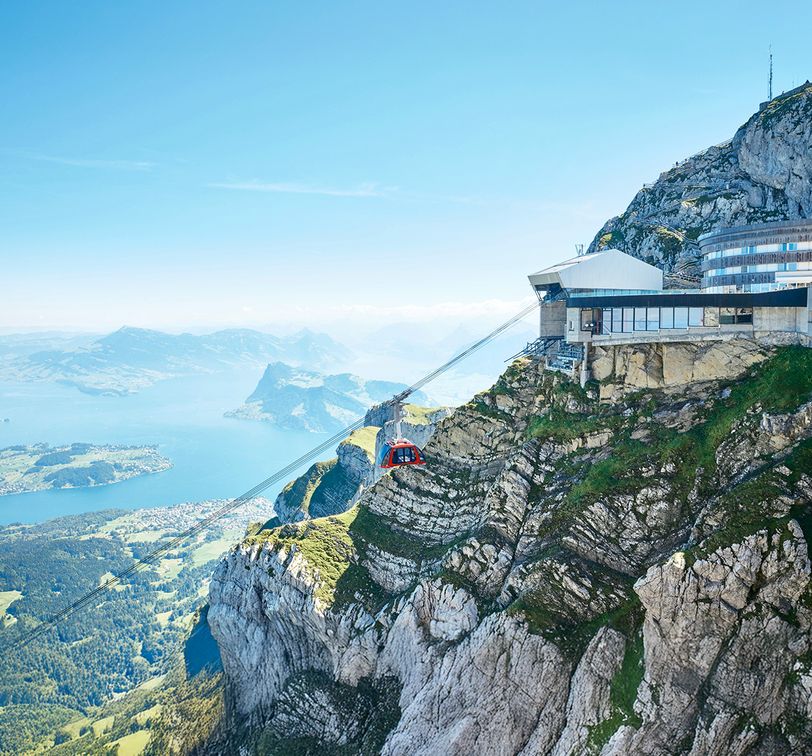 Red cable car ascending a rocky mountain with a station at the top, overlooking a lake and expansive landscape under a clear blue sky.