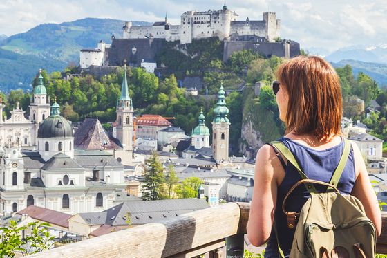 A woman with a backpack overlooks a scenic view of Salzburg, featuring the Hohensalzburg Fortress, historic buildings, and mountains.