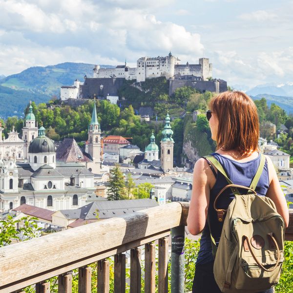 A woman with a backpack overlooks a scenic view of Salzburg, featuring the Hohensalzburg Fortress, historic buildings, and mountains.