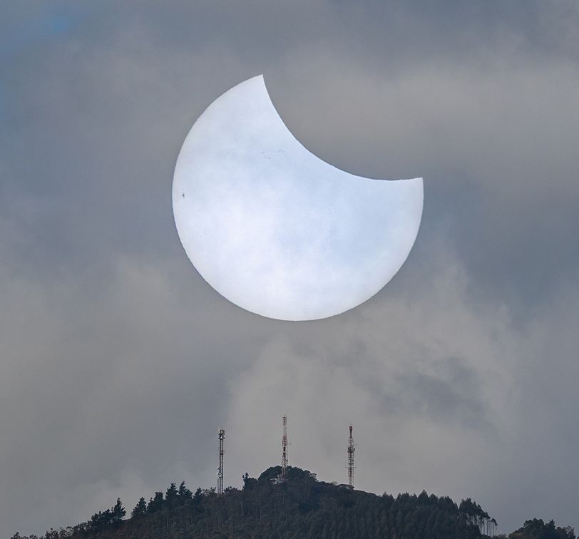 A partially eclipsed sun behind clouds, over a hill with trees and three radio towers.
