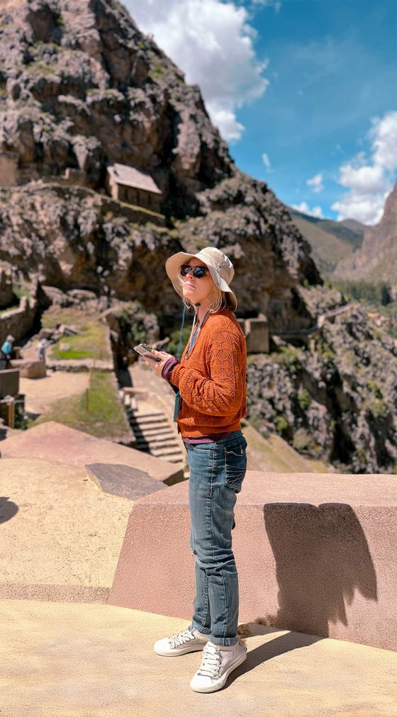 Person in sunglasses and hat stands on mountain path, holding a phone, with rocky terrain and ancient structures in the background.