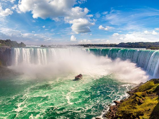 A boat approaches the base of a large, misty waterfall under a bright blue sky with scattered clouds.