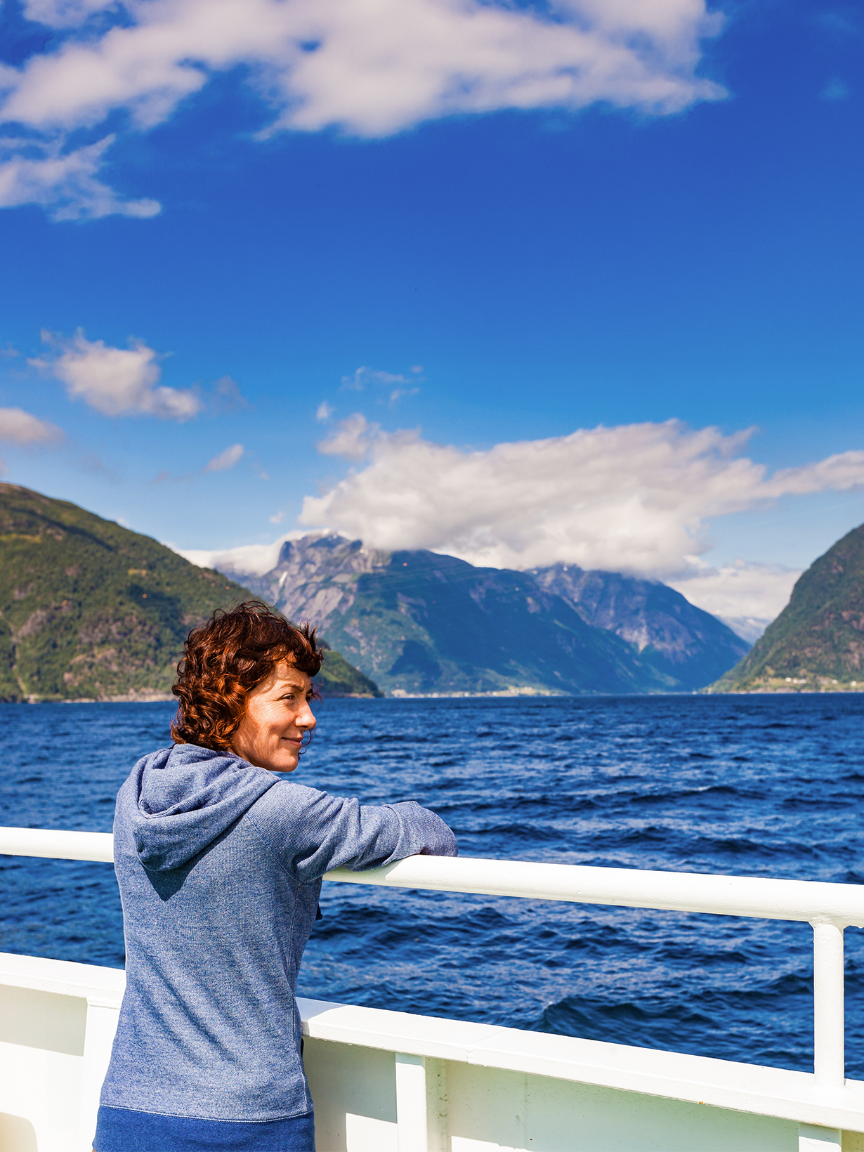 A person in a blue hoodie enjoys a scenic view of mountains and a fjord from a boat, under a bright blue sky with scattered clouds.