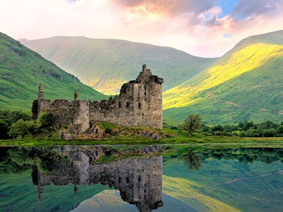 A medieval castle ruin reflected in a calm lake, surrounded by lush green mountains under a dramatic sky.