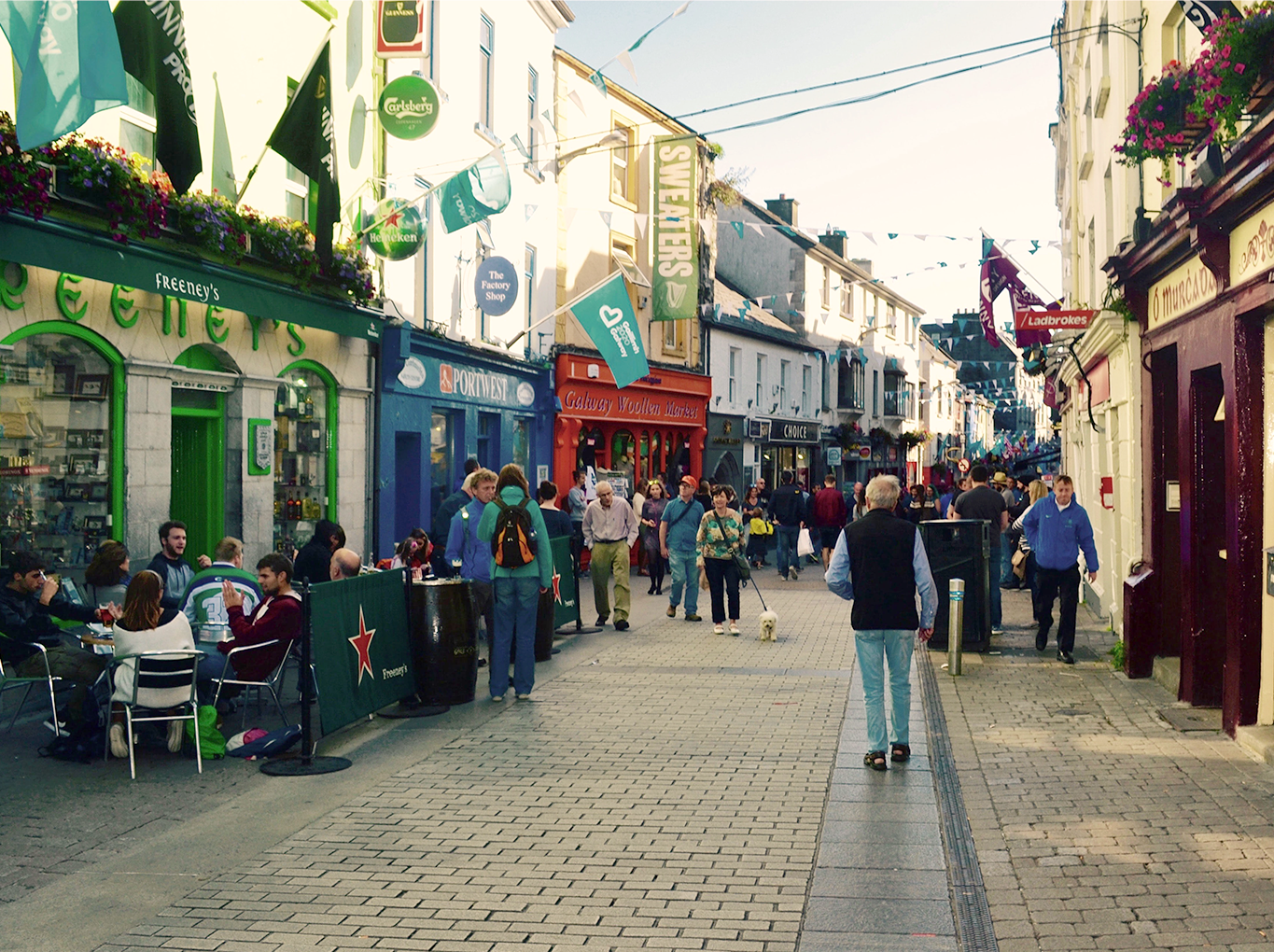 A bustling pedestrian street with people walking and sitting at outdoor cafes, surrounded by colorful shops and hanging signs.