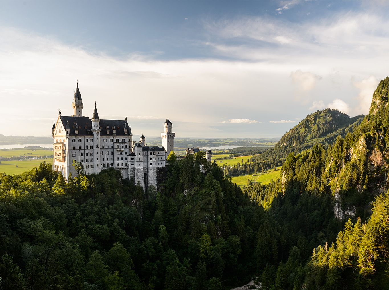 A fairy-tale castle perched on a forested hill, overlooking a lush valley and distant mountains under a partly cloudy sky.