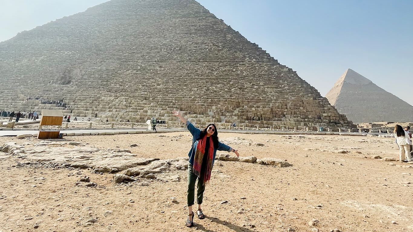 A person joyfully poses in front of the Great Pyramid of Giza, with another pyramid visible in the background under a clear blue sky.
