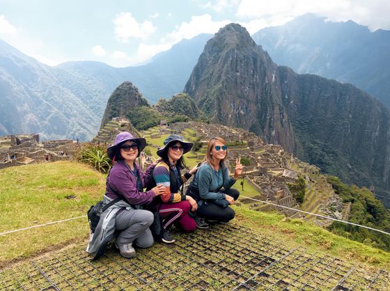 Three people pose in front of the ancient ruins of Machu Picchu with Huayna Picchu mountain in the background under a bright sky.