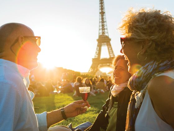 Three people enjoying a sunset picnic near the Eiffel Tower, laughing and holding drinks, with a warm golden glow in the background.