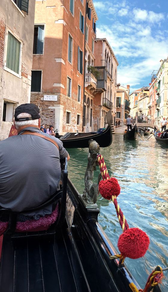 A person rides a gondola on a Venetian canal, surrounded by colorful buildings and other gondolas under a clear blue sky.