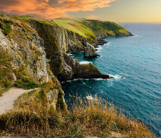 Coastal cliffs with grassy tops overlook a turquoise sea under a vibrant sunset sky, creating a picturesque and serene landscape.