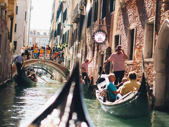 Gondolas navigate a narrow canal in Venice, with people on a bridge above. Sunlight casts shadows on historic brick buildings.