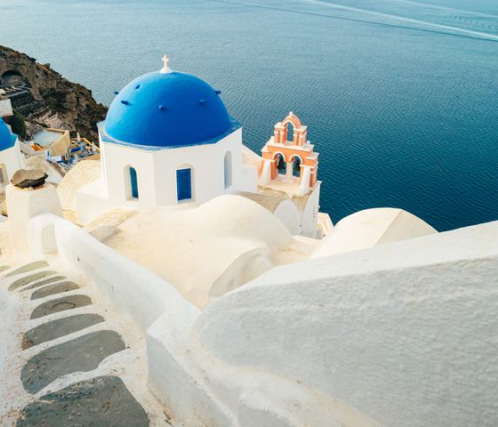 White buildings with blue domes by the sea in Santorini, Greece. A staircase leads down with clear, calm water in the background.