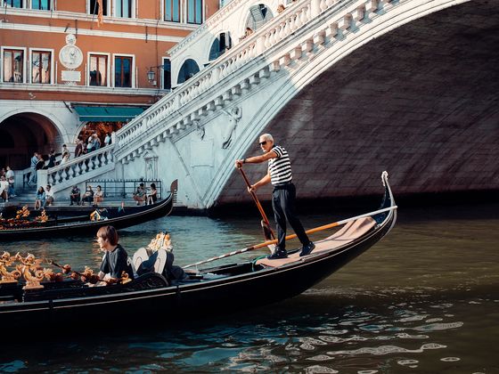 A gondolier rows a gondola on a canal near the Rialto Bridge in Venice, Italy, with a building and tourists visible in the background.