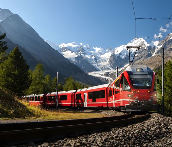 Red train winding through a mountainous landscape with lush green trees and snow-capped peaks under a clear blue sky.
