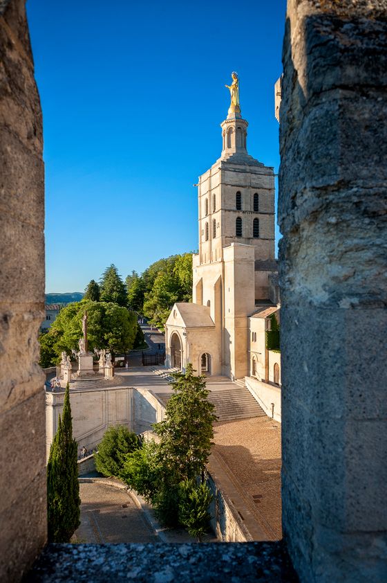 A historic stone tower and church facade viewed through an archway, under a clear blue sky, surrounded by lush greenery.