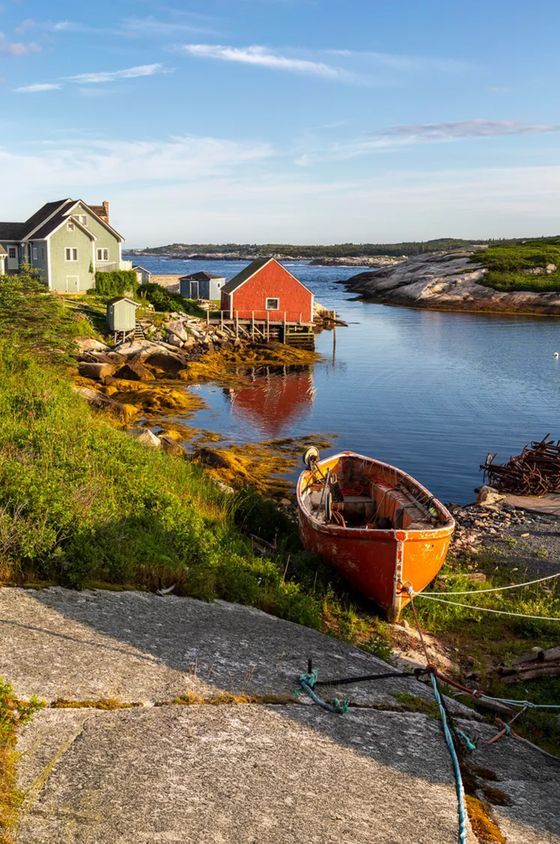Coastal scene with a red boat on grass, a green house, a red boathouse, and calm water reflecting the sky and structures.