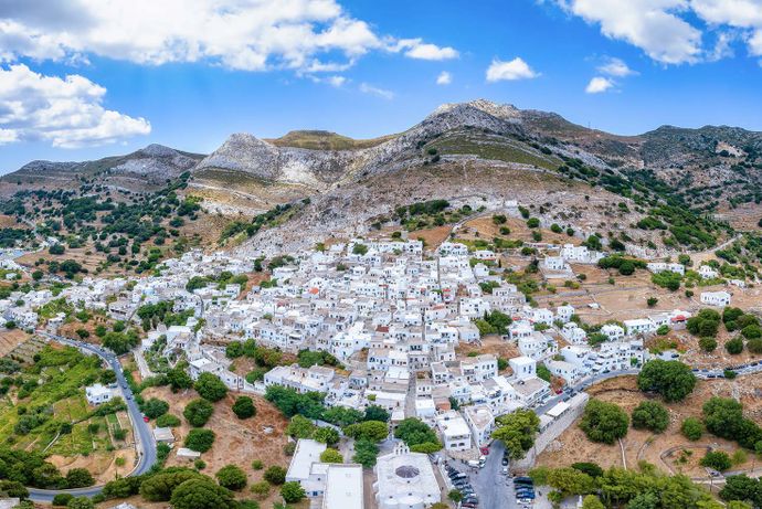 Aerial view of a picturesque hillside village with white buildings, surrounded by greenery and rugged mountains under a blue sky with clouds.