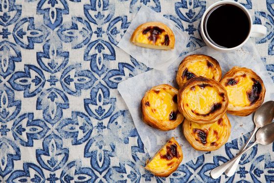 Custard tarts on parchment paper with a cup of coffee and two spoons, set on a blue-patterned tablecloth.
