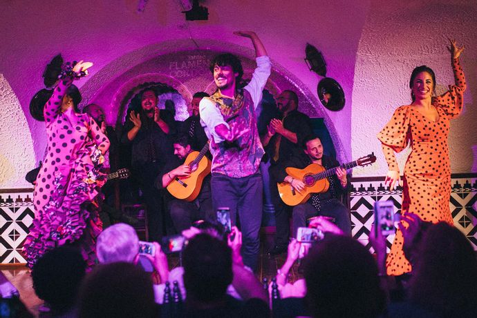 Flamenco dancers perform energetically on stage with guitarists in the background, as an audience captures the moment with their phones.