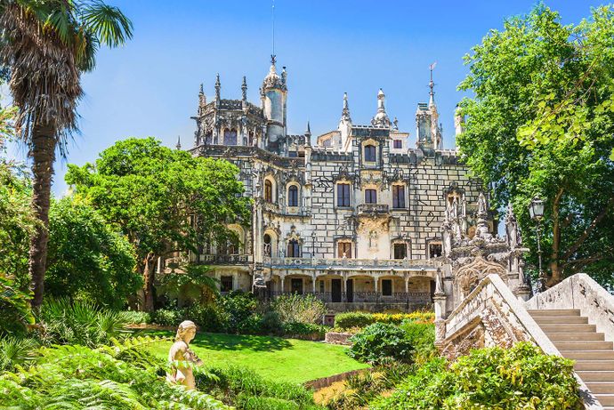 Ornate Gothic Revival palace with intricate details, surrounded by lush greenery, a statue, and stone steps under a clear blue sky.