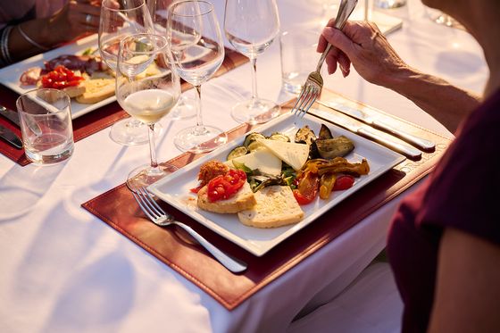 A person holds a fork over a plate of bread, cheese, roasted vegetables, and tomatoes, surrounded by wine glasses on a white tablecloth.